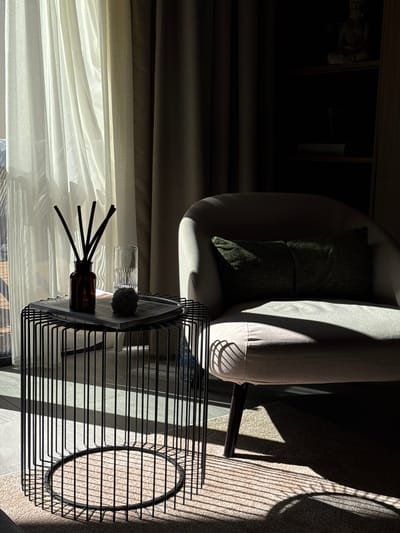 In a dimly lit room, possibly in Bakovići, Montenegro, sunlight streams through sheer curtains, casting dramatic shadows. A plush armchair, covered in a light-colored fabric, sits in the center of the frame, adorned with two dark green cushions. To the left, a minimalist black wire side table holds a glass of water, a brown glass diffuser with black reeds, and a decorative round object. The floor is covered by a light-colored rug with a textured pattern, further enhanced by the play of light and shadow from the side table. The background consists of dark, heavy curtains and a glimpse of a shelf with a small statue, hinting at a tranquil, possibly meditative, atmosphere.