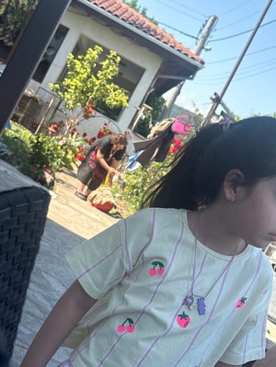The image depicts a daytime scene outdoors, likely in a residential area of Slanchevo, Bulgaria, characterized by a tiled roof house in the background and a clear blue sky. In the foreground, a young girl with dark hair pulled back in a ponytail, wearing a yellow and pink striped t-shirt adorned with fruit patches, is partially visible. She is looking to her right, and appears to be wearing a necklace. Behind her, in the mid-ground, a woman wearing a dark shawl over a patterned dress is tending to plants and a red basin filled with what appears to be laundry or cleaning supplies. The setting includes a lush garden with red flowers and green foliage, suggesting a warm, possibly summer, day. A black woven chair is partially visible on the left side of the frame. The overall impression is of a casual, domestic environment.