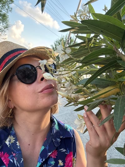 In this close-up, low-angle shot taken outdoors in Alakhadzi, Abkhazia, a person wearing a straw hat with a red and blue striped band is partially visible. The person is also wearing a blue floral-print top and sunglasses. Their right hand, with neatly manicured nails, is reaching up towards a cluster of delicate white flowers and green leaves, suggesting they are interacting with the flora. The background shows a bright blue sky with scattered clouds, and some indistinct trees and possibly power lines, indicating it's daytime with fair weather. The lighting suggests it is likely late afternoon or early morning due to the warm glow on the skin. The overall mood is relaxed and summery, with a focus on the interaction between the person and the natural environment. No visible text is present in the image.