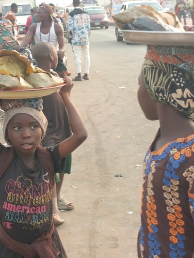 The image depicts a street scene in Alizaga, Nigeria, characterized by pedestrian and vehicular activity. In the foreground, a young dark-complexioned girl is positioned left of center, looking slightly upward and to her left. She wears a dark t-shirt with multicolored "Champion authentic american athletic" text, a brown wrap tied around her waist, and a light-colored head covering. She balances a red-rimmed white tray on her head, laden with multiple items wrapped in leaves or paper, supporting it with her raised right hand. To her right, the upper torso and head of another individual are visible, wearing a brown garment with blue and orange embroidered patterns and a multi-patterned headscarf. This person also carries a metal tray on their head, holding what appears to be a large piece of dried fish or meat and other packaged items.
In the midground, several other individuals are present. A dark-complexioned man in a white tank top and a baseball cap stands near a bicycle, looking left. Further back, another man is seen in a white shirt with a tropical pattern and white trousers. The background contains additional blurry figures and several vehicles, including a white van and two cars on what appears to be a paved road. The ground surface is a worn, light-colored pavement with sparse debris. The scene is illuminated by natural daylight.