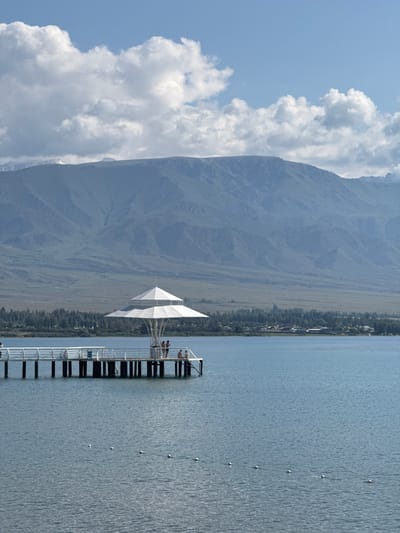 The image shows a serene view of Issyk-Kul lake in Koshkol', Kyrgyzstan. In the foreground, a white pier extends into the calm blue water, with several white buoys visible on the surface. On the pier, a white, umbrella-like structure provides shade, and a few people are gathered beneath it and on the pier, appearing to be relaxing or socializing. Beyond the lake, a line of green trees borders the shore, with a small settlement of buildings visible amongst them. In the background, a large, majestic mountain range dominates the horizon, its slopes rendered in muted tones of green and brown under a bright blue sky filled with fluffy white clouds. The scene conveys a peaceful and picturesque atmosphere characteristic of a lakeside resort town.