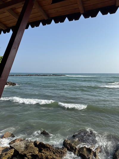 The image presents a view of the ocean from a structure, possibly a beach hut or observation point. To the left, the structure's wooden roof and beam are visible against a clear, bright blue sky. The ocean occupies the majority of the frame. Waves are crashing along a rocky shoreline to the right, with a line of rocks extending into the water. It’s likely that this is in Catia La Mar, Venezuela, due to the rocky shoreline and the style of architecture. No people are visible in the image.