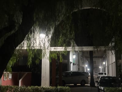 The image depicts an outdoor scene at night, likely in a residential or communal area, possibly a courtyard or parking lot. A large tree with hanging branches is prominent in the foreground, obscuring parts of the background. To the right of the tree, a building with a modern architectural style is visible, featuring a covered walkway supported by pillars. Several parked vehicles are present, including a light-colored SUV or hatchback and a dark-colored sedan. A white van is also visible further back. Exterior lighting illuminates the area, casting a glow on the building and surrounding structures. The ground appears to be paved, and there is dense shrubbery in the lower portion of the image. The overall impression is a dimly lit, quiet night scene. The specific location in Alcàsser, Spain, is not discernible from the visual cues alone.
