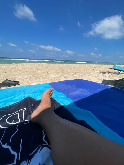 In Ga'ash, Israel, a person is relaxing on a beach. The image is taken from a first-person perspective, focusing on the person's legs and feet as they lie on a towel. The towel is blue and black with a graphic design. Next to the towel are a black sandal and some sand. In the background, the sea and the sky are visible. The sea is a vibrant blue-green with white waves, and the sky is a clear blue with a few scattered white clouds. A beach umbrella and some driftwood are also present in the background. The overall scene depicts a tranquil day at the beach.