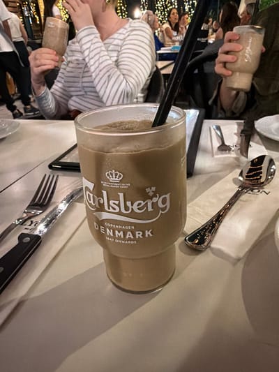 In this close-up shot, a Carlsberg-branded glass filled with a creamy, light brown beverage stands prominently on a white tablecloth. The glass, with its distinctive embossed logo featuring a crown and the text "Carlsberg Copenhagen Denmark 1847 Onwards," is the central focus. A black straw is visible within the drink. To the left of the glass, a silver fork and knife are laid out, partially obscured by the glass. To the right, a silver spoon rests on a white napkin, reflecting the ambient lighting. In the background, out of focus, are people seated at tables, suggesting a restaurant or outdoor dining setting. The overall lighting appears to be dim, with warm tones, possibly from string lights or lamps, creating a relaxed evening ambiance. The image does not provide any specific indicators of the city of Yavne, Israel, or its unique landmarks.