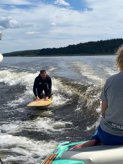 In Gromovo, Russia, a man in a black wetsuit is wakeboarding on a lake. He is on a yellow wakeboard and is in a kneeling position. He is wearing a wetsuit with the number "78" on the chest. The water is choppy and there is a wake behind him. In the foreground, a person wearing a grey t-shirt and blue shorts is sitting on the edge of a boat. The person's feet are visible, and they are bare. The background shows a forested shoreline and a blue sky with white clouds.
