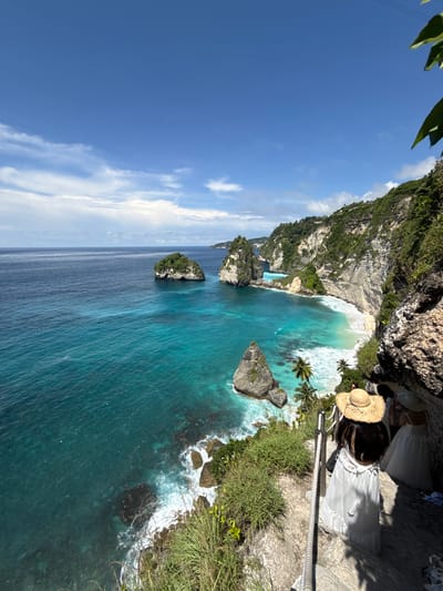 A medium-shot, high-angle, color photograph depicts a panoramic view of the coastline at Pejukutan, Indonesia, featuring cliffs, a beach, the ocean, and a person. The image is rotated 90 degrees counter-clockwise.
In the foreground, a person wearing a white dress and a straw hat stands on a rocky ledge. A safety rope is visible along the edge.
The coastline consists of tall, rugged cliffs covered with green vegetation. A small, white-sand beach nestles at the base of the cliffs. The ocean extends from the beach, transitioning in color from a light turquoise near the shore to a darker blue further out. Several rock formations protrude from the water.
The sky is a clear blue with scattered white clouds. The overall lighting suggests a sunny day.