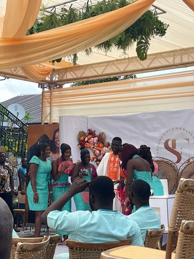 This image depicts an outdoor celebratory event, likely a wedding or engagement, occurring in Kumasi, Ghana. The scene is set under a temporary canopy constructed with metal trusses, draped with translucent orange and cream fabric, and adorned with hanging artificial green foliage including ferns and monstera leaves.
At the center of the image, a group of individuals stands before a white backdrop. The backdrop features floral arrangements in shades of peach, pink, and orange, along with a graphic display reading "SOLOMON AND EMMANUEL" and an intertwined "SE" logo. To the left of the backdrop, a large banner displays a smiling couple.
The central group includes a tall male subject wearing a white robe with prominent orange and white embroidered patterns, possibly a kente design. He is smiling. Facing him is a female subject with reddish-brown hair, wearing a turquoise-colored strapless garment, seen from the back. Three other female subjects stand to their left, all dressed in coordinated turquoise attire. One on the far left wears a short-sleeved dress, smiling. The other two wear two-piece outfits with pink accents; one holds a pink fan, and the other is partially obscured.
To the right of the main group, a small tiered cake in light blue sits on a white pedestal, positioned next to decorative elements made of woven rattan.
In the immediate foreground, two male subjects are seated with their backs to the camera, wearing matching light blue short-sleeved shirts. They occupy woven rattan chairs. Additional guests are partially visible in the left background, one wearing a floral-patterned shirt.
In the upper left background, beyond the event structure, a residential building with a grey corrugated roof and a white satellite dish is visible against an overcast sky.