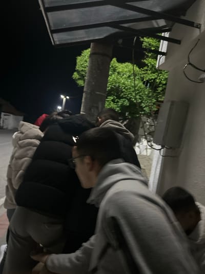 The image captures an outdoor scene at night in Pripek, Bulgaria, featuring a group of people standing in what appears to be a line or close gathering. The perspective is from behind and slightly to the side of the individuals.

Several people, likely young adults, are visible, mostly from the back or side. The person nearest the camera wears a grey hooded sweatshirt and grey pants. To their left, another individual wears a black puffer jacket and spectacles, their head slightly bowed. Further along the line, other people are seen wearing various jackets, including another black puffer jacket with a hint of red, and a light-colored puffer jacket.

The setting suggests an area next to a building, possibly under an overhang or covered walkway. Above the people, a white rectangular electrical fixture or light unit is mounted on a wall, with visible wiring. To the left, a large tree with dense green foliage is prominent, illuminated against the dark night sky. In the deeper background, string lights or small hanging light fixtures add to the ambient illumination. Part of a dark, slatted structure, possibly a fence or part of a canopy, is visible on the far left.

The overall atmosphere is casual, and the individuals appear to be waiting or socializing. The darkness of the sky and the visible artificial lights confirm it is nighttime. The outerwear worn by the people suggests cool weather. There is no visible text in the image.