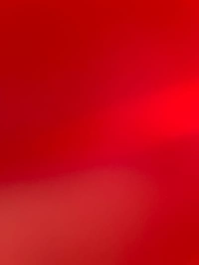 The image is an abstract, out-of-focus depiction of a red background with subtle gradients and highlights. There are no discernible subjects, objects, scenes, or activities taking place. The texture of the image is slightly grainy, with soft light illuminating certain areas, creating a bokeh-like effect. The colors range from a deep, rich red to lighter, almost pinkish hues, particularly towards the right side of the frame. The lighting suggests a bright, possibly artificial light source. No text is visible. The image is a generic, abstract visual that lacks specific context related to Gorno Varshilo, Bulgaria, or any other identifiable location or event.