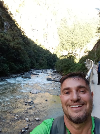 A man is taking a selfie on a bridge in Machupicchu, Peru. He is wearing a green shirt and a backpack, and is smiling at the camera. Behind him is a river flowing through a canyon. The canyon walls are covered in trees and vegetation. Another person is partially visible behind the man on the bridge.