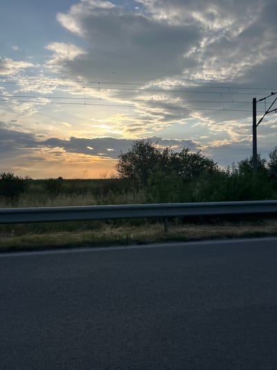 This photograph captures a serene roadside landscape, likely during sunset or sunrise, in Kaloyanovo, Bulgaria. The image is composed with a paved road and a metallic guardrail on the right, suggesting a journey or a view from beside a road.
**Subjects:** The primary subjects include the vast sky, a section of a paved road, a metal guardrail, sparse dry grass, and a line of trees and bushes forming the horizon. A tall utility pole with several power or communication lines stretches across the upper part of the sky.
**Scene and Setting:** The scene depicts a rural or semi-rural environment where human infrastructure (road, guardrail, utility pole) intersects with natural elements. The setting is open countryside, characterized by fields or wild growth alongside the road, leading to a dense treeline in the distance.
**Activity or Event:** No specific human or animal activity is depicted. The image is a still landscape, conveying a moment of natural beauty and tranquility.
**Notable Details:**
* **Time of day:** The most striking detail is the time of day, strongly indicated as either sunset or sunrise. A low sun, not directly visible but casting a brilliant golden-orange glow on the horizon and illuminating the undersides of the clouds, creates a warm and atmospheric light.
* **Weather:** The sky is partly cloudy, featuring a mix of bright, sun-kissed clouds and darker, more voluminous grey clouds, creating a dramatic contrast. The overall weather appears calm.
* **Location cues:** The combination of a paved road, guardrail, and utility lines within a natural, unurbanized setting points to a countryside or rural highway location. The dry, yellowish grass suggests a particular season, possibly late summer or autumn.
* **Emotions:** The scene evokes feelings of peace, contemplation, and appreciation for the natural world and the changing light of the day.
* **Visible text:** There is no visible text within the image.