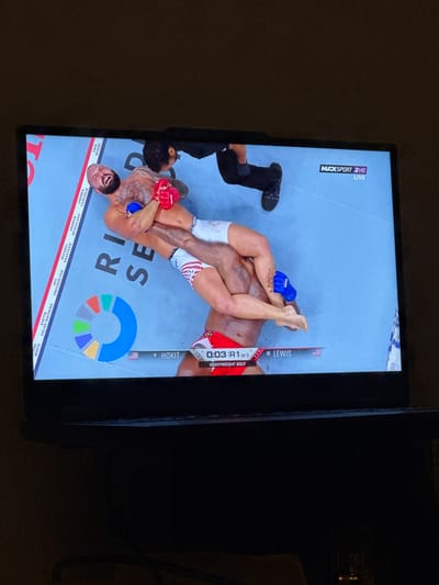 The image shows a television screen displaying a mixed martial arts (MMA) fight. Two male fighters are grappling on a blue mat, with one fighter appearing to be in control. Text overlays indicate fighter names, round information, and broadcast details. The screen is positioned on a black surface, likely a table or desk, in a dimly lit room. No specific location in Enschede, Netherlands, can be identified.