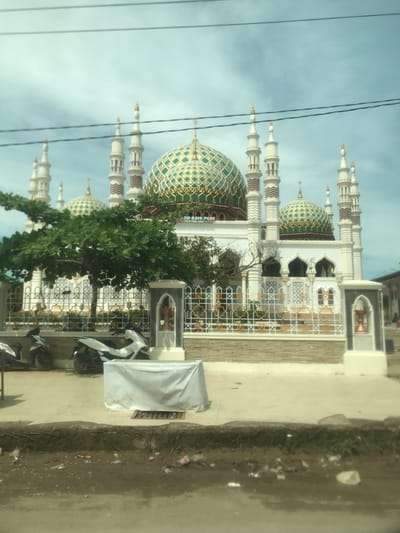 The vertically oriented image, captured during the daytime in Kotapantonlabu, Indonesia, presents a detailed view of a mosque. The mosque stands out with its pristine white structure, featuring multiple slender minarets topped with finials and adorned with distinct brown and white chevron patterns. Several large domes, beautifully tiled in an intricate green and gold diamond pattern, crown the mosque.
In the midground, a vibrant green tree partially screens the lower section of the mosque, while ornate white fencing with gold geometric designs borders the mosque's premises. Parked in front of the fence are several motorbikes, including one distinctively white and black vehicle, and another covered by a white tarp next to a white rectangular unit. Overhead, two dark electrical wires traverse the light blue, partly cloudy sky.
The foreground shows an unpaved strip of ground, littered with some small debris, running alongside a paved area. A visible banner or sign affixed to the mosque structure clearly displays the text "JAYA PASE" and "PANTUNLABU," providing a specific location cue. The overall scene is bright, devoid of people, and highlights the architectural beauty of the mosque against a clear sky.