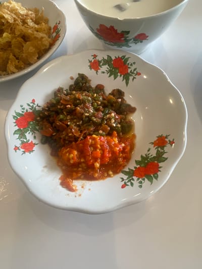In a brightly lit room, likely a dining area in Medan, Indonesia, a white ceramic plate with a floral design is presented on a white tabletop. The plate contains two mounds of chopped chili, one green and one red, glistening with oil. To the left of this plate is another white ceramic bowl filled with what appears to be fried food. In the background, a portion of a white bowl with similar floral accents is visible, containing a light-colored liquid, possibly a beverage or soup. The overall scene suggests a meal is being served or enjoyed.