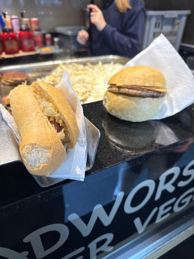 The image captures a close-up view of a food stall counter, likely within a market or event setting in Sint-Niklaas, Belgium. In the foreground, on the left, a hot dog-style sandwich, containing a sausage and a quantity of what appears to be sauerkraut or sautéed onions, is presented in a metal serving holder lined with white paper. To the immediate right, a separate round bread roll, containing a meat patty and a thin slice of melted cheese or bacon, rests on the black, reflective countertop, partially enveloped in white paper. In the background, a large, rectangular metal griddle holds a substantial amount of thinly sliced, sautéed onions, from which faint steam is visible. To the left of the griddle, stacked round, flat objects are discernable, consistent with prepared patties or bread. Behind the counter, a person wearing a dark blue long-sleeved top is partially visible, with hands engaged in handling food preparation utensils. The front of the counter below the serving surface features a black panel with white, stylized lettering, partially displaying "WORS" and "ER VEG", indicative of a vendor specializing in sausage and potentially vegetarian burger products.