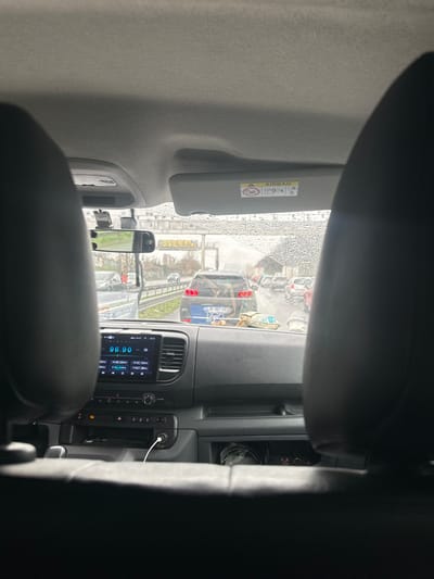 The image presents a forward-facing view from the rear passenger compartment of a vehicle in Aulnay-sous-Bois, France, observing a traffic scene under rainy conditions. The perspective is framed by the dark headrests of the front seats. Above, the vehicle's grey headliner is visible, along with a deployed passenger-side sun visor displaying an "AIRBAG" warning label.

The dashboard, primarily black, features an infotainment system showing "96.90" as a radio frequency and the time "15:13." A white charging cable is connected to a power outlet below the infotainment unit. A rearview mirror is visible, with a small decorative charm hanging from it.

Beyond the rain-splattered windshield, a multi-lane road is congested with vehicular traffic. Directly ahead is a dark blue SUV or crossover, displaying illuminated red taillights and what appears to be a graphic or sticker on its rear. Other vehicles are visible in the distance, indicating traffic slowdown or stoppage. To the left of the road, a metal guardrail separates the roadway from an adjacent lower-level area. An elevated concrete overpass spans the width of the road in the mid-ground, featuring visible graffiti, including the letters "YEL," on its supporting pillar. Bare trees and urban structures are present in the distant background under overcast skies. No individuals are explicitly discernible within the frame.
