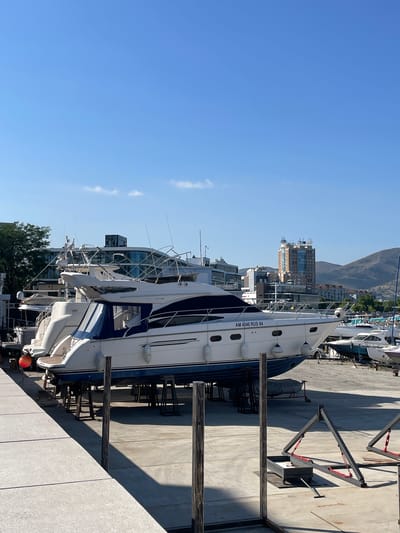 A yacht is docked on a sunny day. The yacht is white and blue, with the registration number "AM 4346 RUS 64" visible on its side. It is supported by wooden stands on a concrete dock. Other yachts and buildings are visible in the background. The sky is clear and blue, indicating fair weather. The scene appears to be a marina or harbor. The time of day is likely daytime, given the bright sunlight and shadows. No people are visible. The location is identified as Maiorovskii, Russia.