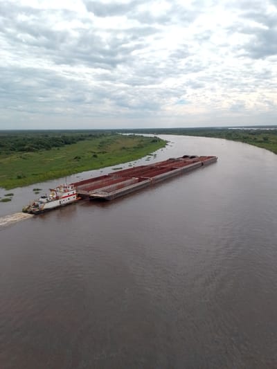 An aerial view depicts a river scene, presumably in the city of Concepción, Paraguay. A long barge, composed of connected sections, is being pushed by a tugboat. The barge is dark brown or red in color. The tugboat is white with red stripes and appears to be actively navigating the river. The river itself is wide and murky brown. The river banks are lined with lush, green vegetation, transitioning into a dense forest in the background. The sky is overcast with a layer of white clouds.