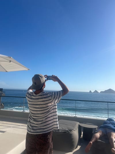 The image shows a man on a balcony in Cabo San Lucas, Mexico, taking a picture with his phone. The balcony overlooks the ocean and a rocky coastline. The man is wearing a striped shirt, patterned shorts, and a baseball cap. Another person is lying on a lounger in the background. A white umbrella is partially visible in the foreground. The sky is clear and blue.