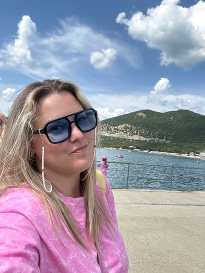 A close-up, eye-level outdoor shot captures a blond woman, likely in her twenties or thirties, positioned slightly right of center, looking directly at the camera with a slight smile. She is wearing black-framed, rectangular sunglasses with blue-tinted lenses, secured by a delicate white and pink beaded glasses chain. A small silver stud piercing is visible on her left nostril. She wears a pink, long-sleeved shirt with a white, marbled or tie-dye pattern. Her hair is long and blond, parted on the side. To her immediate left, a portion of another person's hand with light pink nail polish is visible.

Behind the woman, a tranquil blue-green body of water, likely the Black Sea, extends to a distant shoreline. A dark metal railing separates the concrete foreground, where the woman stands, from the water. On the far left of the midground, a young child wearing a pink hat and brightly patterned clothing stands by the railing, looking towards the water. In the midground water, a small red inflatable boat with two occupants is visible. The shoreline features a long, narrow beach populated with numerous small, brightly colored beach umbrellas and loungers. Several white buildings or structures are scattered along the beach.

In the background, a large, verdant mountain covered in dense green foliage rises prominently from the shoreline, with a lighter, rockier section visible on its lower slopes. The sky above is a clear blue with scattered white cumulus clouds, indicating a bright and sunny day. The scene is set in Bol'shoi Utrish, Russia.