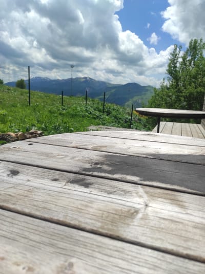 The image depicts a close-up view of a weathered wooden table in the foreground, with a blurred background of mountains and a cloudy sky. A portion of another wooden table and bench are visible to the right. The background reveals a grassy hillside with a fence and some trees. The location appears to be a scenic overlook, possibly in Gudauri, Georgia, given the mountainous terrain. The sky is partly cloudy with patches of blue. There are no people or animals visible in the frame.