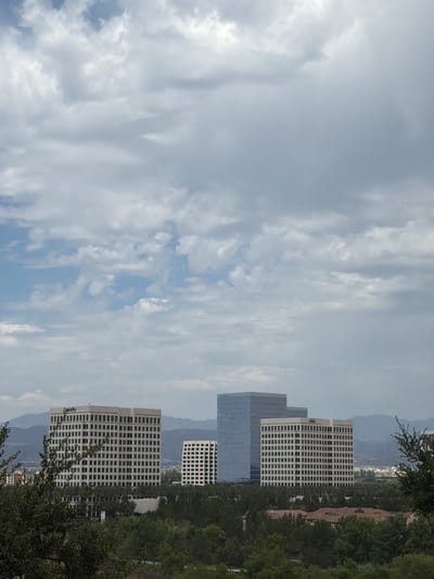 This image depicts a series of modern office buildings in Irvine, California. The buildings are tall and rectangular with numerous windows, suggesting a business or corporate setting. They are surrounded by lush green trees and foliage, indicating a well-maintained, possibly suburban or park-like environment. In the background, distant mountains are visible under a cloudy sky. The sky is predominantly overcast with large, wispy clouds, suggesting a moderate or slightly humid day. There are no people visible in the image. The lighting is diffused due to the cloud cover, implying it is likely daytime, but the specific time is not clear. One of the buildings has the word "Generality" visible on its side. The overall scene conveys a sense of order and professional activity.