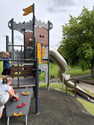 A children's playground in a park, likely within the city of Wakefield, United Kingdom, features a large, castle-themed play structure with a silver tube slide. Two young boys are visible on the structure; one in a blue shirt and black shorts is climbing, and another in a grey shirt and light grey shorts, wearing noise-canceling headphones, is near the climbing wall. The ground cover is a mix of dark rubber mulch and green artificial turf. In the background, a grassy field is dotted with people picnicking and walking. A mature tree with lush green foliage stands to the right of the frame. The sky is overcast with clouds.