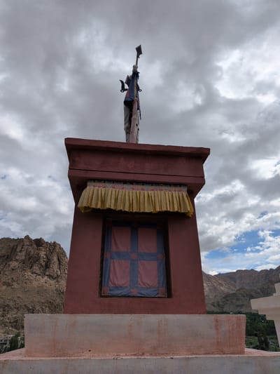 A low-angle, wide shot captures a Buddhist stupa in Phyang, India, against a backdrop of a cloudy sky and rugged mountains. The stupa is a multi-tiered structure with a reddish-brown facade. A yellow, pleated fabric hangs above a square window that is adorned with a blue and pink checkered pattern. Atop the stupa, a person, dressed in a light-colored shirt and pants, stands holding a flag with a triangular black emblem at its top. The person's body is partially obscured by the flag pole and a blue and red cloth. The stupa is positioned on a light-colored base, and in the background, the arid, mountainous terrain of Ladakh can be seen.