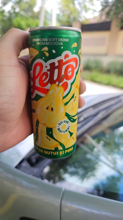 A person's hand is holding a green and yellow can of "Latto" sparkling soft drink from Moldova. The can features images of sliced pears and the text "CU SUCI NATURAL" and "DE GUTUI ȘI PERE". The background shows out-of-focus greenery and a building, suggesting an outdoor urban setting. The image appears to have been taken in Rîșcani, Moldova.