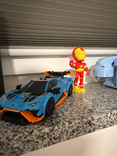 The image shows toys on a speckled countertop. A blue and orange toy car with a black spoiler is on the left. To the right of the car, a small red and yellow figure resembling Iron Man is standing, with its left hand raised. Behind Iron Man, a light blue toy building is partially visible. The countertop has a granite-like pattern. In the background, horizontal blinds are visible against a white wall. The image appears to be taken indoors, with no indication of the city of Sankt Augustin, Germany.