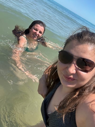 Two women are in the ocean at Shkorpilovtsi, Bulgaria. The woman in the foreground is wearing sunglasses and a dark-patterned swimsuit, and is smiling at the camera. The woman in the background, wearing a green swimsuit, is also smiling and looking at the camera as she swims. The water is a light green hue, and the sky is a clear blue.