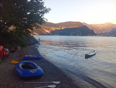 In Hergiswil (NW), Switzerland, on a pebble beach, an inflatable blue boat labeled "200" rests on the shore. Nearby, another blue inflatable boat and a yellow object are also on the beach. Two paddles lie on the ground in front of the boats. In the water, a person stands near the shore, and a paddleboard floats further out. The background features a serene lake with mountains rising in the distance, their slopes dotted with buildings. The sky is clear and bright, with the warm glow of the setting sun illuminating the mountain peaks.