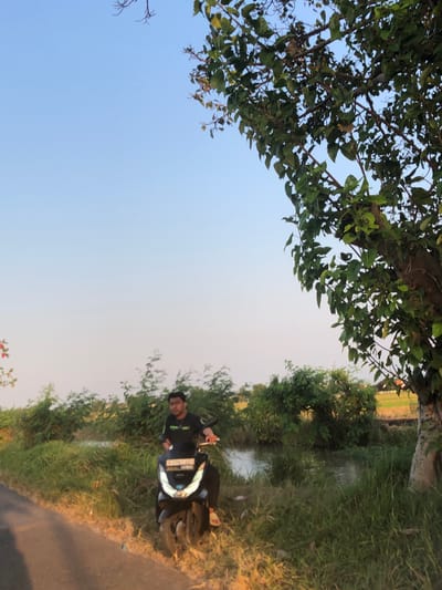 In Keboguyang, Indonesia, a young man is seated on a black motorcycle parked on the side of a dirt road. He is wearing a black t-shirt and dark pants. The motorcycle's headlight is illuminated. The road is bordered by tall green grass and a small body of water. In the background, lush green foliage and trees are visible under a clear blue sky. The overall scene depicts a rural landscape during daylight.