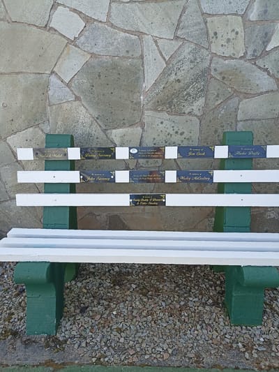 The image shows a white bench with green concrete supports situated outdoors. The bench has multiple plaques attached to its backrest, indicating memorial inscriptions. The plaques are in gold and black and bear names and phrases such as "In memory of Patrick Walsh," "In memory of Dinny Sweeney," "In memory of Jim Cook," "In memory of Hudie Duffy," "In memory of John Sweeney," "In memory of Wesley McCartney," and "In memory of Paddy Buddy O'Donnell & Eddie Sharkey." There is also a plaque with more text that is difficult to read in full. The background is a stone wall. The ground in front of the bench is covered in gravel. The scene is consistent with a public memorial bench located in Loughanure, Ireland.