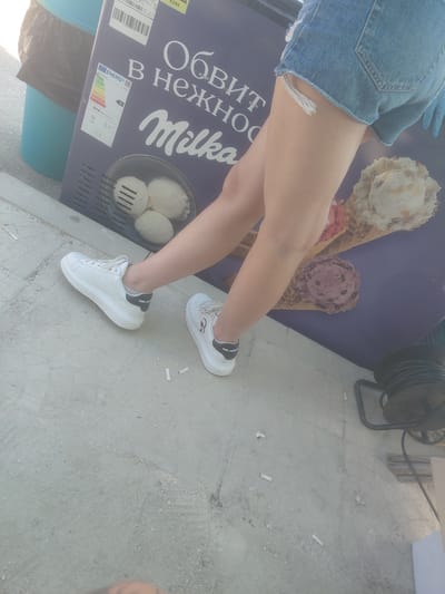 This image captures an outdoor scene in the city of Kostievo, Bulgaria, featuring the lower body of a person, a commercial ice cream display, and surrounding ground details.
The central subject is the lower torso and legs of an individual, dressed in light blue denim shorts with frayed edges and white platform sneakers. The left sneaker clearly displays "KARL LAGERFELD" branding on the black heel accent. The person is standing with their feet slightly apart, angled towards the left.
Adjacent to the person is a large, upright commercial ice cream freezer or display unit, predominantly purple in color. The unit features prominent white Cyrillic text, including the brand name "Milka" in its characteristic script font. Additional Cyrillic text, partially visible, reads "ОБИТ В НЕЖНО..." (Obit v Nezhno...), suggesting a phrase like "Coated in tender...". An energy efficiency label is affixed to the unit, displaying an energy class 'D' and indicating '949 KWh/annum'. Illustrated on the display are various ice cream items: three scoops of white ice cream in a metal container, and two large ice cream cones, one appearing to be vanilla or caramel flavored and the other a darker, possibly chocolate or berry, flavor.
The ground is a light gray concrete surface, appearing somewhat rough and uneven. Scattered across the concrete are several small, white cylindrical objects, identified as cigarette filter tips. In the upper left background, a light blue plastic bin containing a black trash bag is partially visible. To the far right, a black power cord reel is situated on the concrete, with electrical cords visible. The scene is brightly illuminated, suggesting daytime.