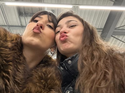 Two women pose closely for a selfie, making kissy faces at the camera. The image is oriented such that they appear upside down. The woman on the left wears a black leather jacket and has curly hair. The woman on the right wears a brown fur coat and large hoop earrings. The background appears to be a commercial space with overhead lighting and metal beams.