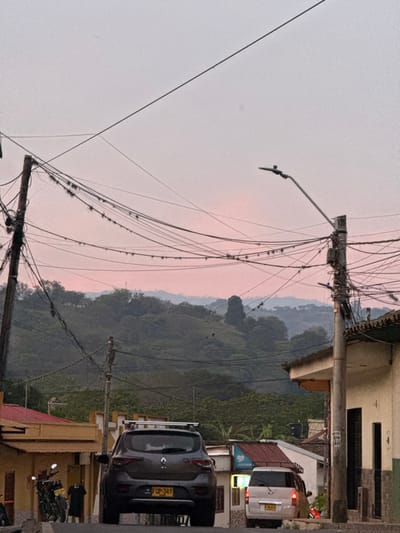 The image presents a street scene in Timaná, Colombia, captured during twilight hours, indicated by the pale pink and light grey sky. The view looks uphill along a paved street.

In the immediate foreground and midground, numerous black electrical and communication cables crisscross the upper portion of the frame, originating from and connecting between two prominent utility poles. The left pole, dark in color, is densely covered with these cables, some of which exhibit small, green epiphytic plant growth. The right pole, lighter in color, also bears a substantial number of wrapped cables and features a curved streetlamp arm extending leftward with a light fixture.

Centrally positioned on the upward-sloping street is a dark grey SUV, possibly a Renault, parked facing away from the viewer. It is equipped with a silver roof rack and displays Colombian license plate "FDP-349." To the left of the SUV, a single-story building with a red-orange tiled roof and a yellow-painted facade is partially visible. Two motorcycles, one black, are parked on the street near this building. A dark-clad individual is observed standing or walking with their back to the camera adjacent to the motorcycles.

On the right side of the street, a light-colored building with a dark roof and a beige facade, featuring a dark doorway, is visible. Further up the street on the right, a light-colored minivan or small SUV is either stationary or in slow motion, displaying license plate "XCM-656." An adjacent building shows an illuminated interior or storefront.

The background consists of multiple layers of verdant, tree-covered hills and mountains, appearing somewhat hazy and blending into the pinkish horizon.