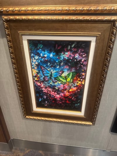 A framed painting of two butterflies is mounted on a textured gray wall. The painting features a vibrant, impressionistic background with various colors like blue, pink, purple, green, and yellow. The butterflies are depicted with clear outlines and bright colors; one is predominantly blue with pink and black markings, and the other is green with black markings. The frame of the painting is ornate and golden, with a ribbed texture on its outer edges. Below the painting, a metallic strip runs along the bottom of the wall, and a patterned carpet is visible on the floor. The overall scene suggests an indoor setting, possibly a public space or a home. There is no indication of the city of Motril, Spain in the image.