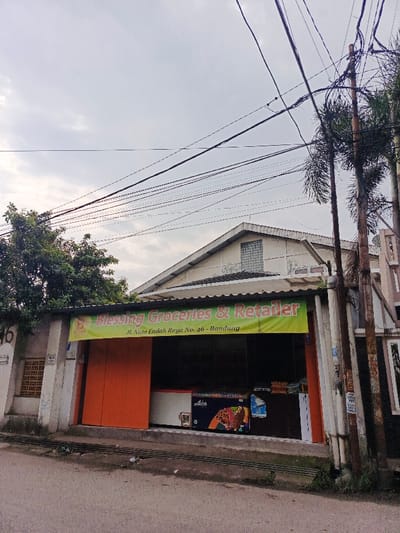 A single-story shopfront with a green awning labeled "Blessing Groceries & Retailer," located at Jl. Nata Endah Raya No. 46 in Bandung. An orange door is on the left, while the main shopfront is open, revealing freezers with "Aice" branding. A white building with a gabled roof stands behind the shop. Overhead are tangled power lines, and tall trees frame the sides of the image.