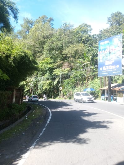 This scene is located in Ka Thaung (Upper), Myanmar.
A curved, two-lane asphalt road winds through a densely vegetated, hilly environment under a clear blue sky. Bright sunlight casts sharp tree shadows onto the road surface. In the foreground, a white four-door SUV is moving from left to right. In the mid-ground, a silver four-door car and two motorcycles, each with a single rider (one wearing a white helmet), are proceeding away from the viewer in the opposing lane.
The left side of the road is bordered by a low, weathered brick wall and a narrow concrete drainage ditch, behind which dense bamboo and other green foliage rise steeply. The right side of the road features a thick jungle-like forest with utility poles and overhead wires running alongside. A large, rectangular billboard stands prominently on the right, displaying an advertisement with the word "Garden," text indicating "5,1 Km," and an address: "Jl. Jend. Sudirman, Kab. Cianjur". Beyond the billboard, a small, open-fronted roadside stall with a light brown roof and a blue and white striped umbrella is visible, accompanied by a few chairs. A yellow diamond-shaped road sign indicating a right curve is partially visible among the foliage on the right side.
