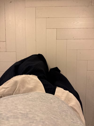 The image displays a high-angle view of a person's lower torso and legs, standing on a light-colored wooden floor in Gondomar, Portugal. The floor consists of light beige or off-white wooden planks arranged in a staggered pattern, showing minor surface wear. The person is wearing multiple layers of clothing: a light grey, textured garment (possibly a sweatshirt) in the foreground, covering the abdomen; beneath it, a light beige or cream-colored fabric, likely a shirt or trousers, with visible folds; and a darker, possibly navy blue, pinstriped fabric (likely trousers) visible on the outer sides of the legs. No faces, hands, or distinct features of the individual are visible.
