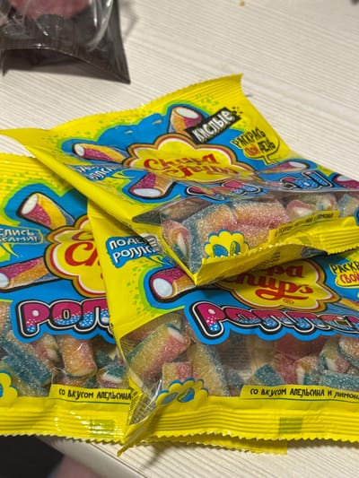 This is a close-up, eye-level photograph of a bag of gummy candies, likely taken indoors on a light-colored, wood-grain surface. The bag is primarily yellow and blue, with prominent pink and red lettering spelling out "Роллс" (Rolls). The candies themselves are visible through the clear plastic packaging and appear to be cylindrical, with multiple colors including pink, yellow, and blue, dusted with sugar.
Several pieces of text are visible on the packaging. On the yellow sections, in Russian, phrases like "Кислые" (Sour), "РАСКРАСЬ" (Paint/Color), "ПОДЕ" (Something like "under"), and "САМИ!" (Yourselves!) can be deciphered. There are also phrases indicating flavor, such as "СО ВКУСОМ АПЕЛЬСИНА И ЛИМОН" (With the taste of orange and lemon) and "СО ВКУСОМ АПЕЛЬСИНАХА ЛИМОНА" (which appears to be a variation or misspelling of the same flavor indication). The phrase "PACKE" is also visible.
The lighting suggests an indoor setting, possibly a home or store, with no discernible time of day or weather cues. The focus is sharply on the candy bag, with the background surface slightly out of focus. There are no people visible in the image, and the overall mood is commercial and focused on the product. The presence of Russian text indicates a possible geographical context of Russia, specifically Gamovo, Russia, as per the location context provided.