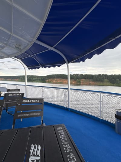 The image shows a view from the deck of a boat on a river, possibly in Zuevo, Russia. The deck is covered by a blue awning supported by white poles. Several black tables and chairs are arranged on the blue carpeted deck. The chairs have the word "БАЛТИКА" (Baltika) written on their backs. The river flows past the boat, with a tree-covered embankment on the opposite side. The sky is overcast. There are no people visible in the image.