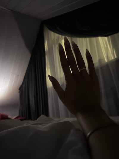 A low-light, high-angle shot shows a person's left hand, palm facing outward, against a backdrop of sheer curtains illuminated by an unseen light source. The hand is adorned with a ring on the ring finger and a bracelet on the wrist. The bed linens in the foreground are soft and rumpled, suggesting a relaxed or intimate setting. The scene is framed by dark fabric and a white wall with a textured ceiling, possibly in a bedroom. The location is identified as Doma 1096 Km, Russia.