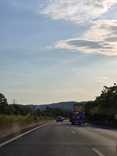 The image depicts a stretch of highway near Dudelange, Luxembourg. The viewpoint is from within a vehicle, likely a car, travelling on the right-hand lane of a multi-lane motorway.
To the right of the roadway is a concrete barrier and beyond it is greenery suggesting a hillside. Power lines are visible in the distance.
In the roadway there is a vehicle with a large chevron board and blue arrow pointing to the left, presumably directing traffic. Several other vehicles are also on the road.
The sky is partly cloudy with the clouds concentrated on the left. The overall light suggests either early morning or late afternoon.