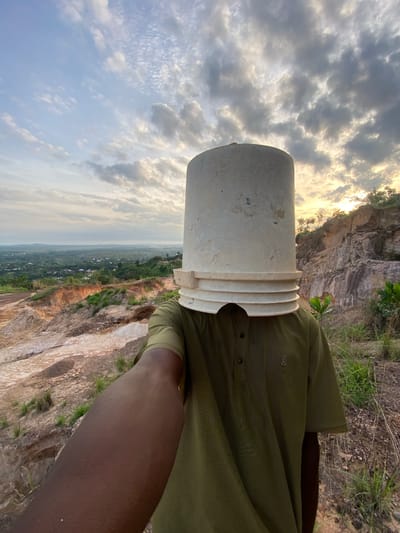 A person stands on a rocky cliff edge in Ufueseni, Nigeria, looking out over a vast landscape. A white plastic bucket is playfully placed upside down over their head. The person is wearing a dark olive green, horizontally striped short-sleeved shirt, and their dark-skinned left arm is visible in the foreground. The bucket is plain white with distinct concentric ridges around its open rim, which rests on the person's head.
The setting reveals a dramatic natural environment. In the immediate vicinity of the person are rugged, exposed light-colored rock formations, suggesting a quarry or natural erosion. Sparse green grass and small bushes grow in patches on the rocky ground. Beyond the cliff, a sweeping vista unfolds, featuring a wide valley or plain covered in lush green vegetation. Numerous small structures, indicative of a village or scattered settlements, are visible in the distance, extending towards the hazy horizon where faint hills or mountains can be discerned.
The sky is partly cloudy, with a mix of grey and white clouds. A warm, golden light, emanating from the setting or rising sun, illuminates the clouds and the distant landscape, creating soft shadows and a serene atmosphere. The overall scene captures a moment of playful interaction with a grand natural backdrop in West Africa.