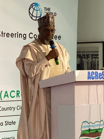 A man is speaking at a podium. He is wearing a patterned cap and light pink robes. He is holding a microphone with a blue and green handle. The podium has the word "ACRes" written on it vertically in blue letters. The background behind the man contains the text "THE WORLD Steering Committee" and a globe icon. The text "(ACR Country Cli a State nigeria" is also visible near the bottom of the image. A framed picture hangs on the wall behind the podium. The location is identified as Kwarau, Nigeria.