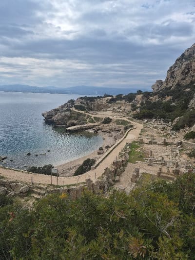 An elevated wide-angle photograph captures an archaeological site along a rocky coastline under an overcast sky in the Municipal Unit of Loutraki - Perachora, Greece. The foreground consists of dense green foliage with scattered yellow blossoms.
The sky is uniformly covered with thick, grey clouds, indicating overcast conditions. To the left, a calm, dark blue-green body of water meets a rugged shoreline featuring a small, crescent-shaped cove with a light-colored beach. A winding, unpaved path follows the curve of the coastline, running parallel to the water. A rectangular, dark-colored sign on a pole is visible beside this path near the cove.
Extensive stone archaeological ruins are visible to the right of the path, spreading across a rocky, undulating landscape. These consist of numerous foundations, scattered blocks, and low-lying structural remnants of ancient buildings. Sparse green vegetation, including shrubs and bushes, is interspersed throughout the ruins and on the rocky terrain. The right side of the image is dominated by a steep, rugged rock face or cliff.
In the far background, across the water, distant mountains or hills are faintly discernible on the horizon, with some indistinct coastal development visible along the shoreline. No individuals or active elements are present in the scene. The lighting is diffused and even, characteristic of a cloudy day.