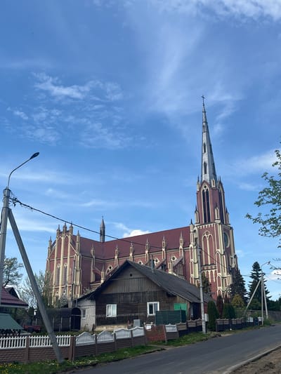 The image shows the Church of the Holy Trinity in Гервяцкі сельскі Савет, Belarus, captured from a low angle. The church, constructed of red brick with light-colored trim, features a tall, pointed steeple. Next to the church, there is a weathered wooden house with a dark roof. A road runs along the right side of the image. A utility pole with a street light and associated wires are in the foreground, partially obscuring the view. The sky is blue with scattered clouds. The surrounding area includes grass, fencing, and some trees.